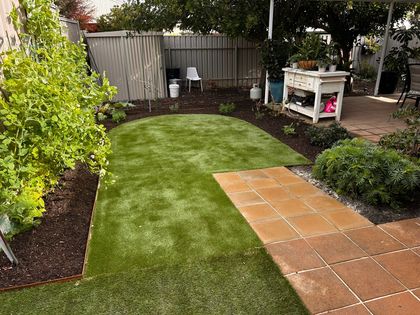 Outdoor landscaped walkway with curved yellow planter, wet pavement, fence posts, and a tree under blue sky