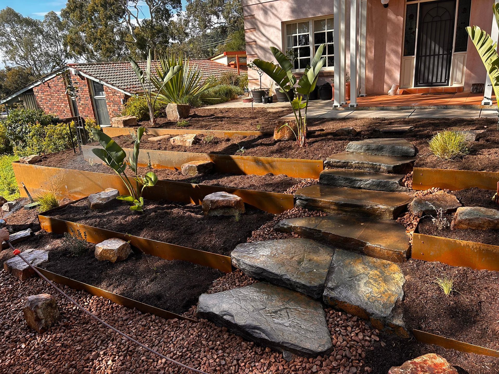 Curved garden bed with young shrubs and dark mulch beside a green lawn and brick house wall