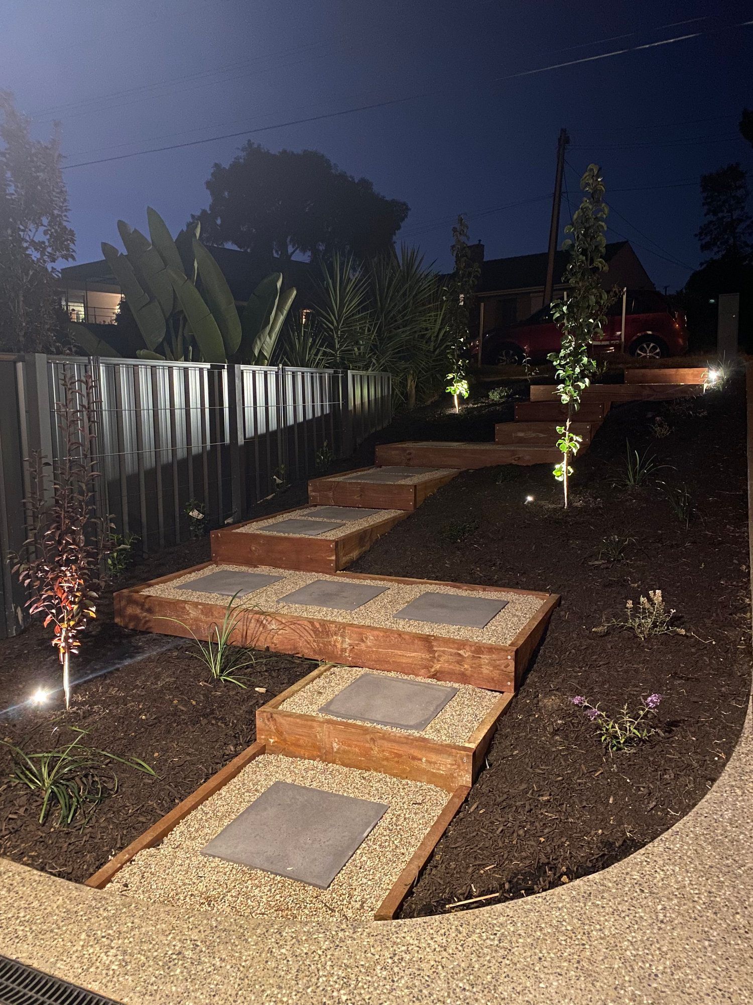 Nighttime garden pathway with stepping stones, wooden steps, and small landscape lights along a fence. Nighttime garden pathway with stepping stones, wooden steps, and small landscape lights along a fence.