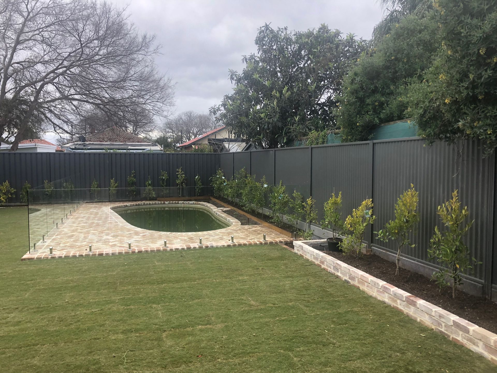 Backyard with a small in-ground pool, lawn, stone patio, and tall fence under cloudy skies