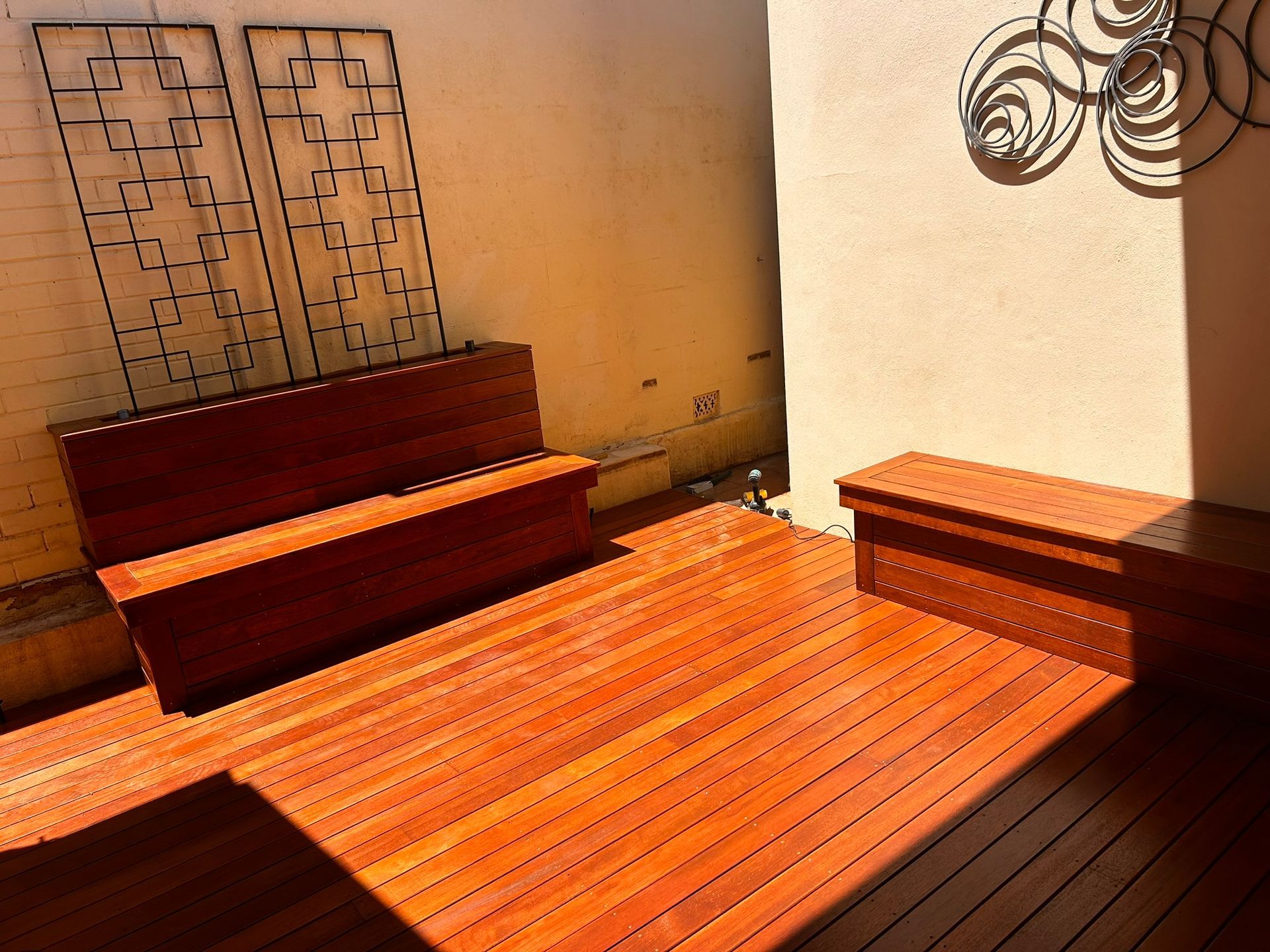 Covered backyard patio with wooden built-in benches, planter boxes, and a fenced perimeter under shade cloth.
