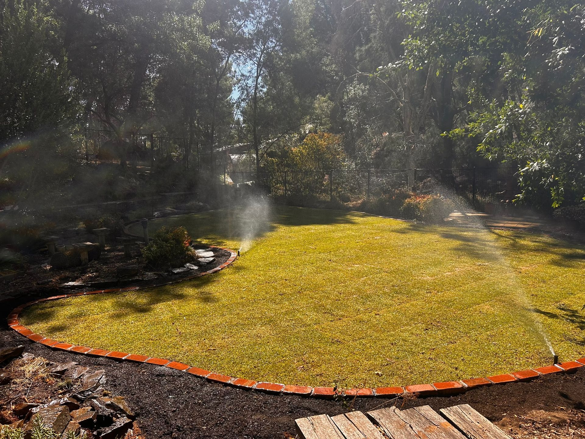Yellow circular lawn spraying water in a shaded garden, bordered by orange edging.