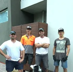 Four men standing outdoors in front of a building, with a dog at their feet. Four men standing outdoors in front of a building, with a dog at their feet.