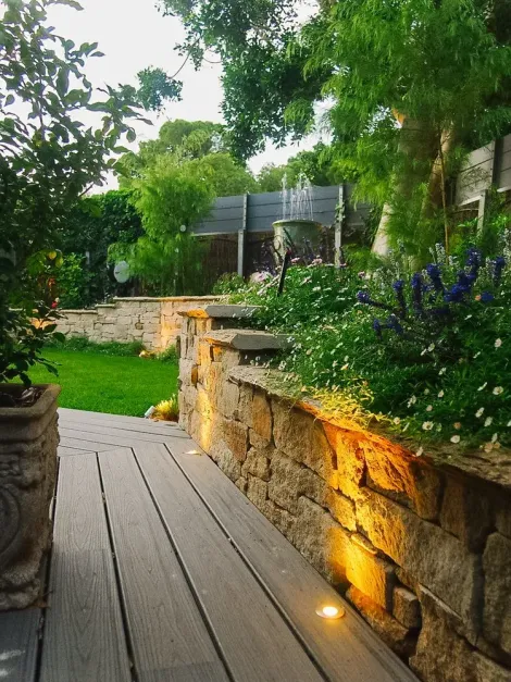 Garden walkway with stone wall lighting beside lush greenery and wooden deck steps Garden walkway with stone wall lighting beside lush greenery and wooden deck steps