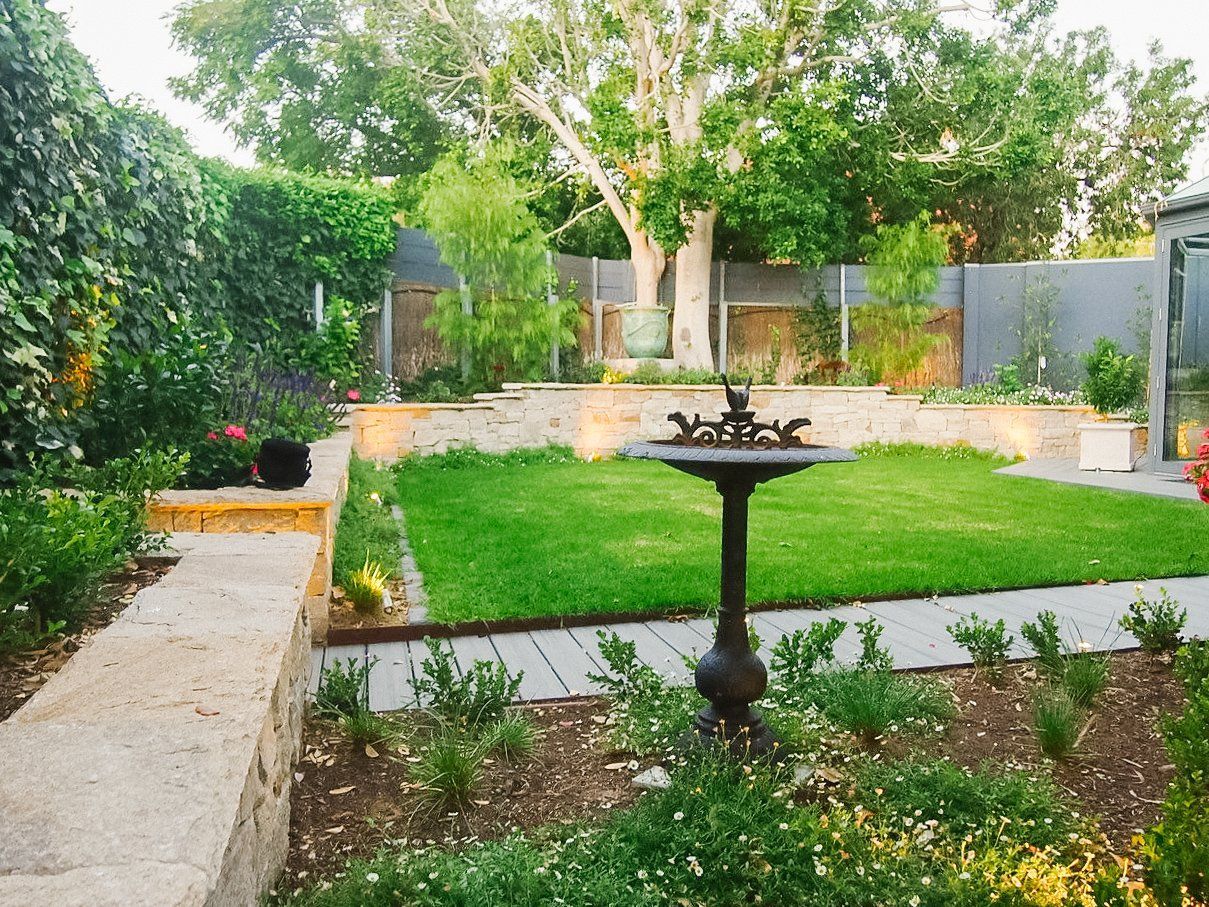 Lush backyard garden with green lawn, stone paths, shrubs, and a central birdbath or fountain. Lush backyard garden with green lawn, stone paths, shrubs, and a central birdbath or fountain.