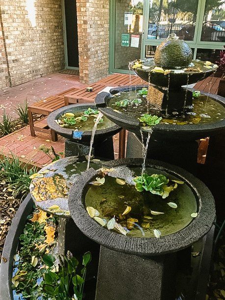 Three black outdoor fountains with water lilies beside a brick patio and wall Three black outdoor fountains with water lilies beside a brick patio and wall