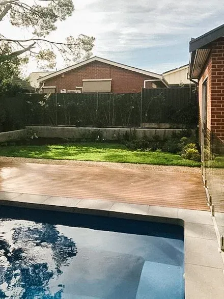 Backyard pool beside a brick patio, green lawn, and tall fence under a cloudy sky
