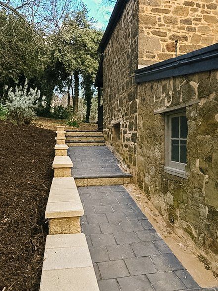 Stone walkway beside a rustic building, with steps, low wall, and trees along a sloped path Stone walkway beside a rustic building, with steps, low wall, and trees along a sloped path