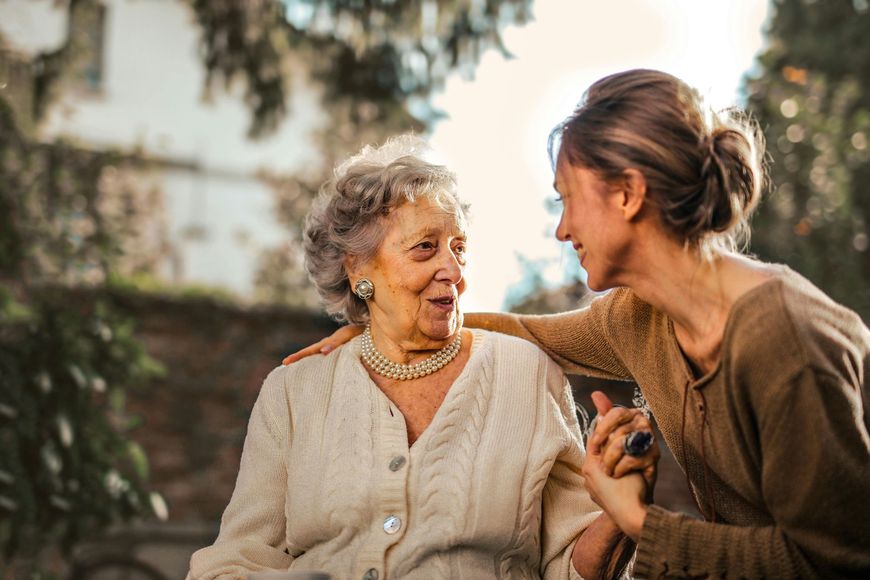 Woman in brown top supports elderly woman in white sweater, outdoor setting.