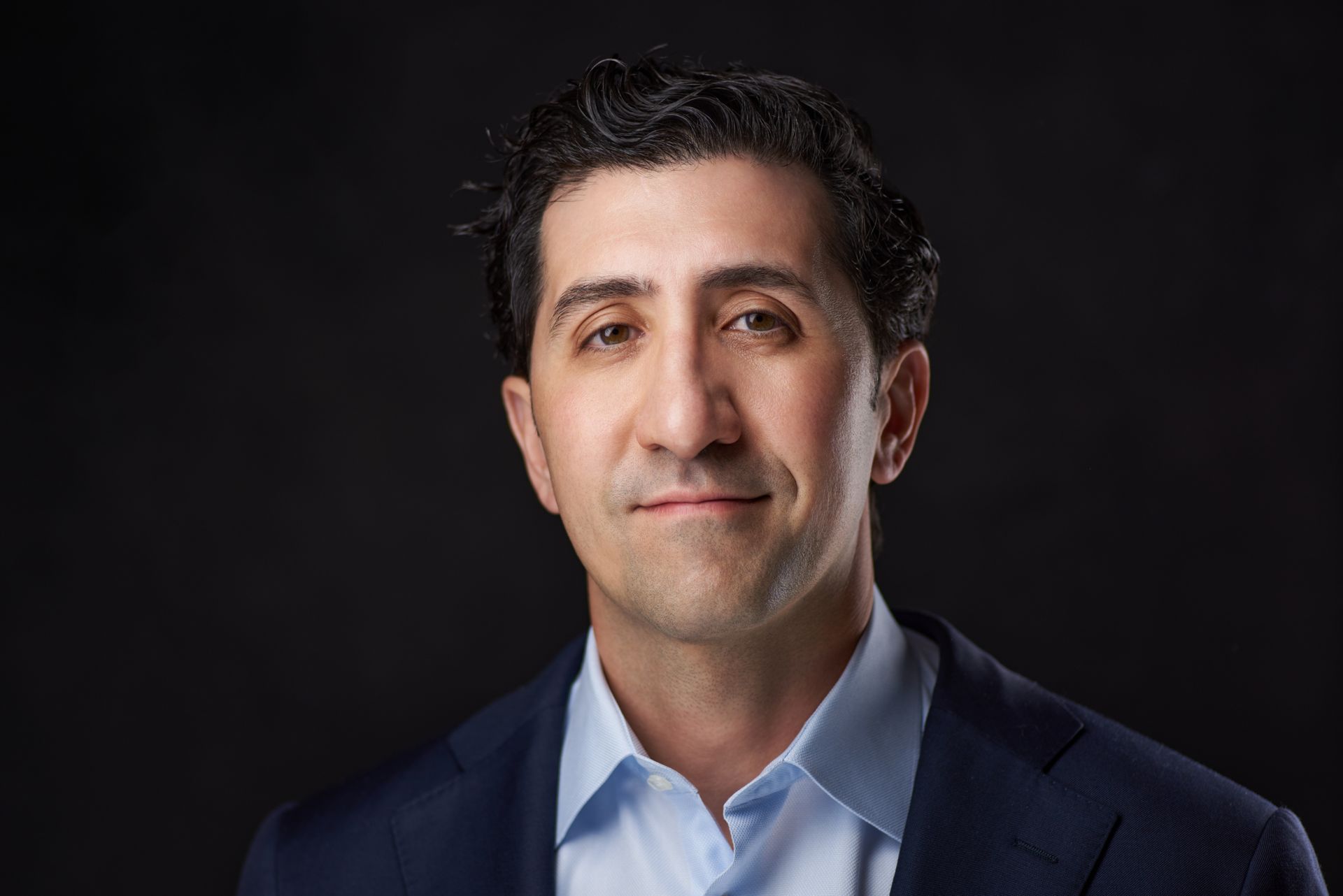 Professional headshot of a man in a navy suit against a dark studio background.