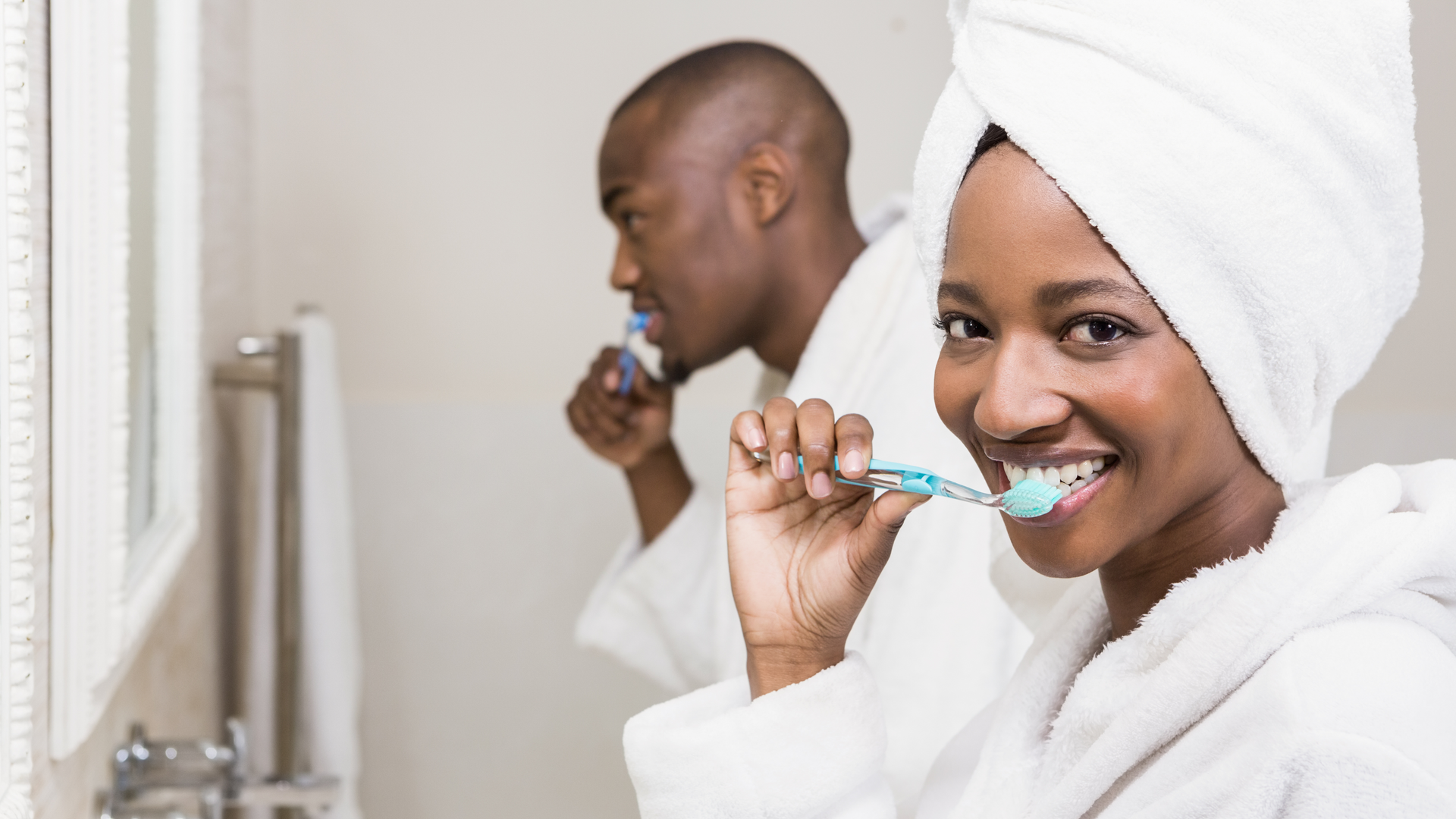 A African American couple in the bathroom brushing their teeth together in their white bathrobes.