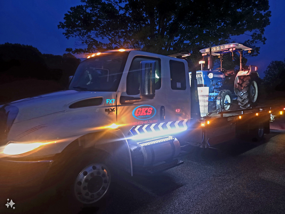 A tow truck with a tractor on the back is parked on the side of the road at night.