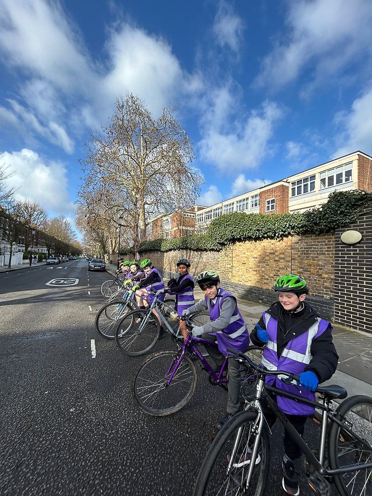 Cyclists in purple jackets and helmets line up on a tree-lined street under a bright blue sky.