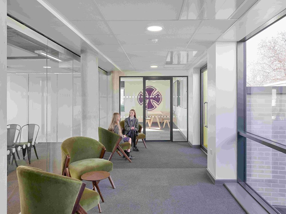 Bright office lounge with green chairs, glass walls, and two people sitting near a window.