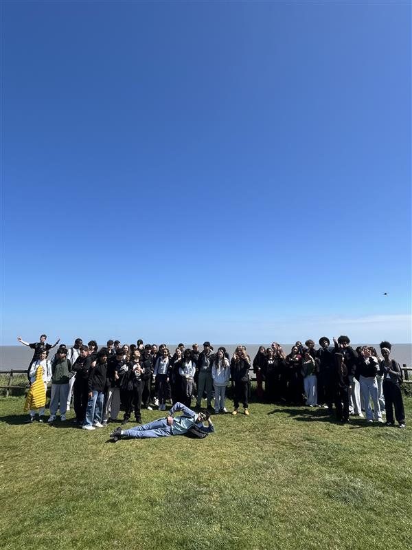 Crowd of people standing on a grassy field under a clear blue sky, with a few blankets laid out in front.