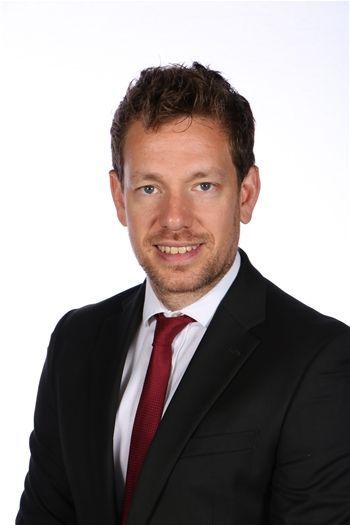 Porträt eines Mannes in schwarzem Anzug, weißem Hemd und roter Krawatte vor weißem Hintergrund.