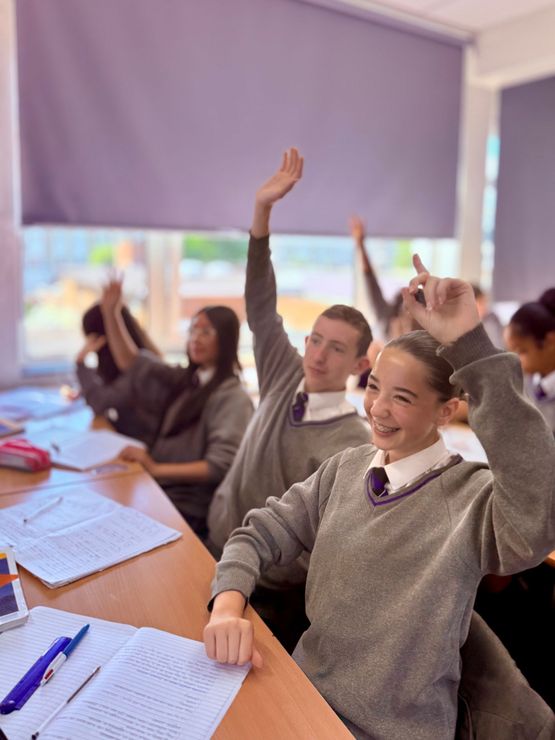 Schüler in grauen Schuluniformen sitzen mit erhobenen Händen an ihren Tischen in einem Klassenzimmer.