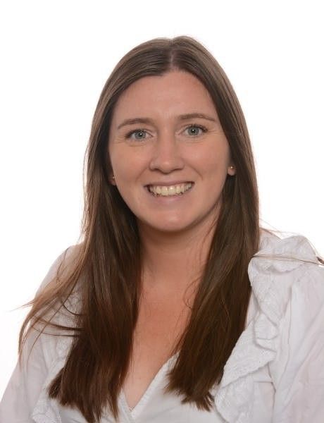 Lächelnde Person in Blazer und weißer Bluse vor hellem Hintergrund