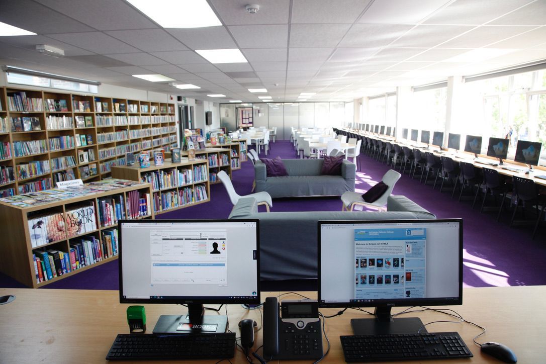 Computerraum der Bibliothek mit zwei Monitoren, Bücherregalen und lila Sitzgelegenheiten in einem hellen Raum