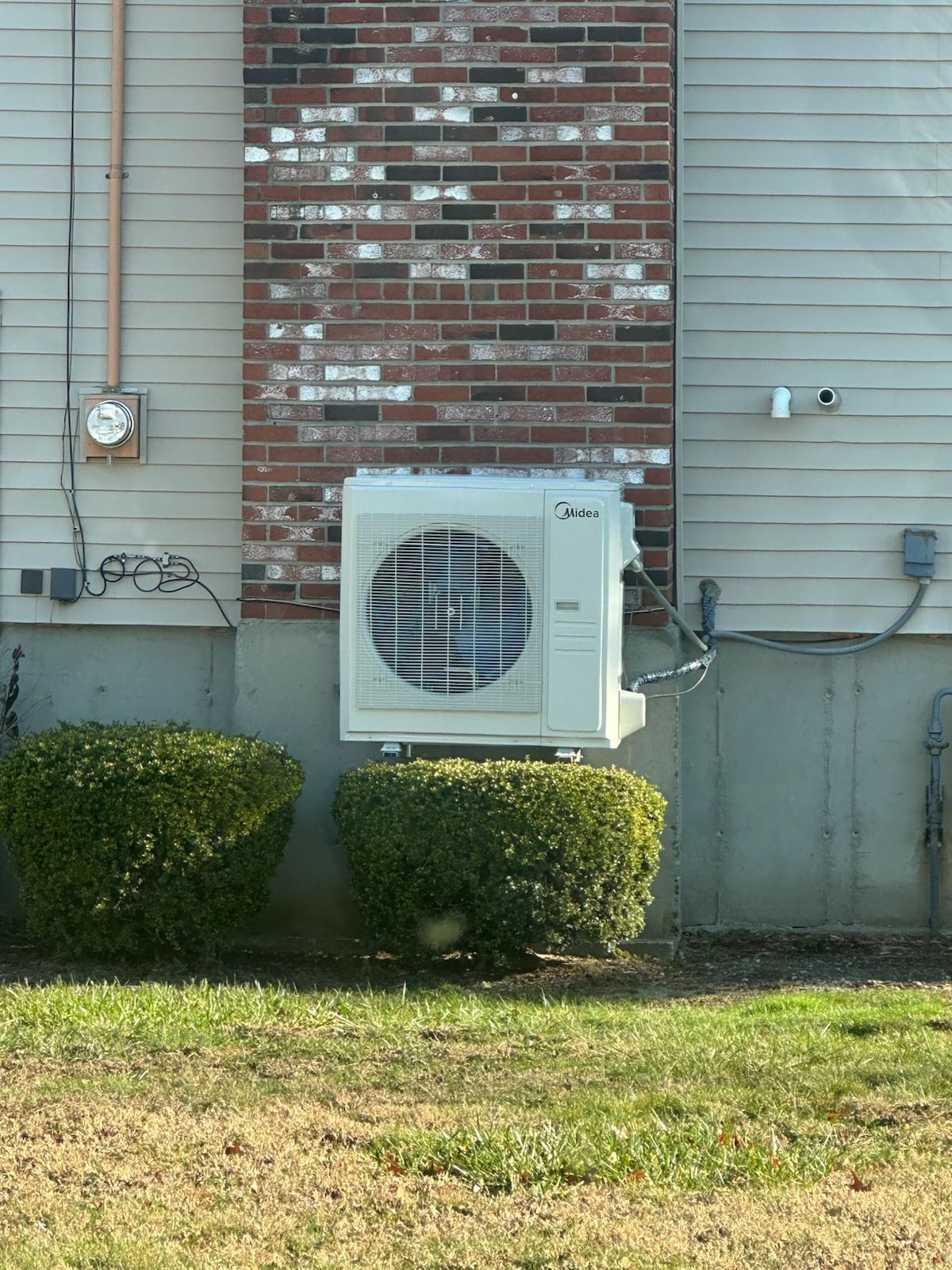 Outdoor HVAC heat pump mounted on a brick and siding wall, with bushes and grass below.