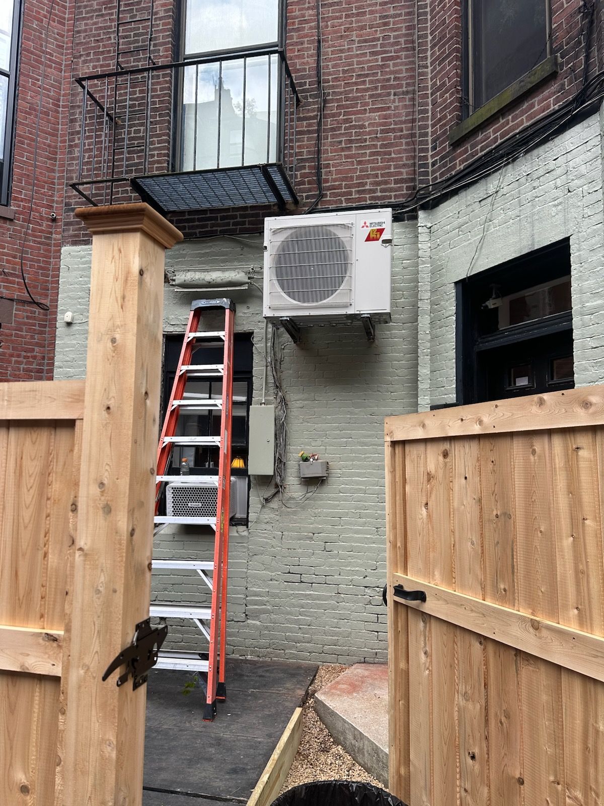 Ladder leaned against brick building beside air conditioner and wooden fence in narrow alley