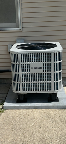 Outdoor air conditioning unit beside a house wall, with a black fan top and white grille.