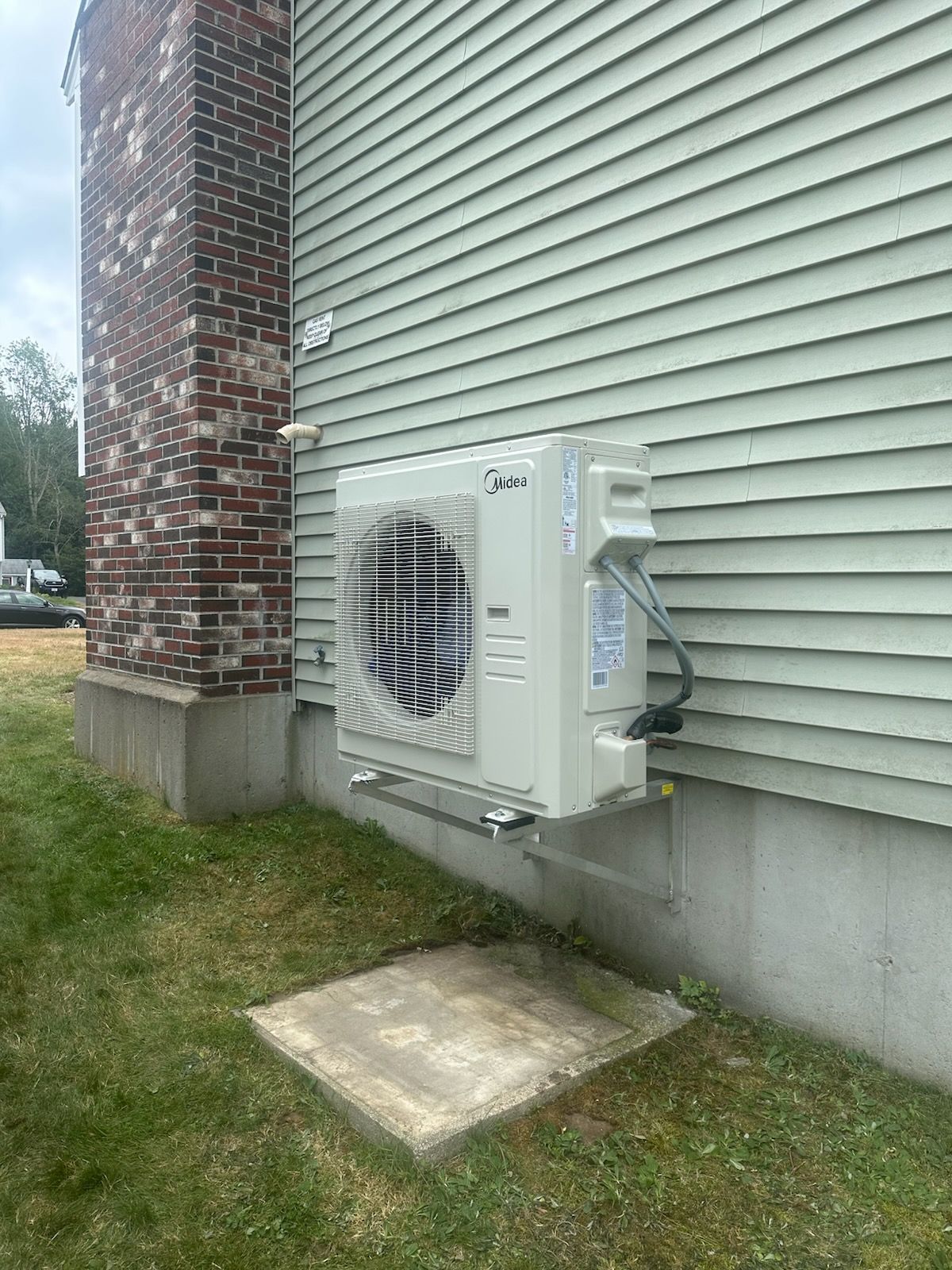 Outdoor air conditioner unit mounted on house siding beside a brick chimney and concrete slab
