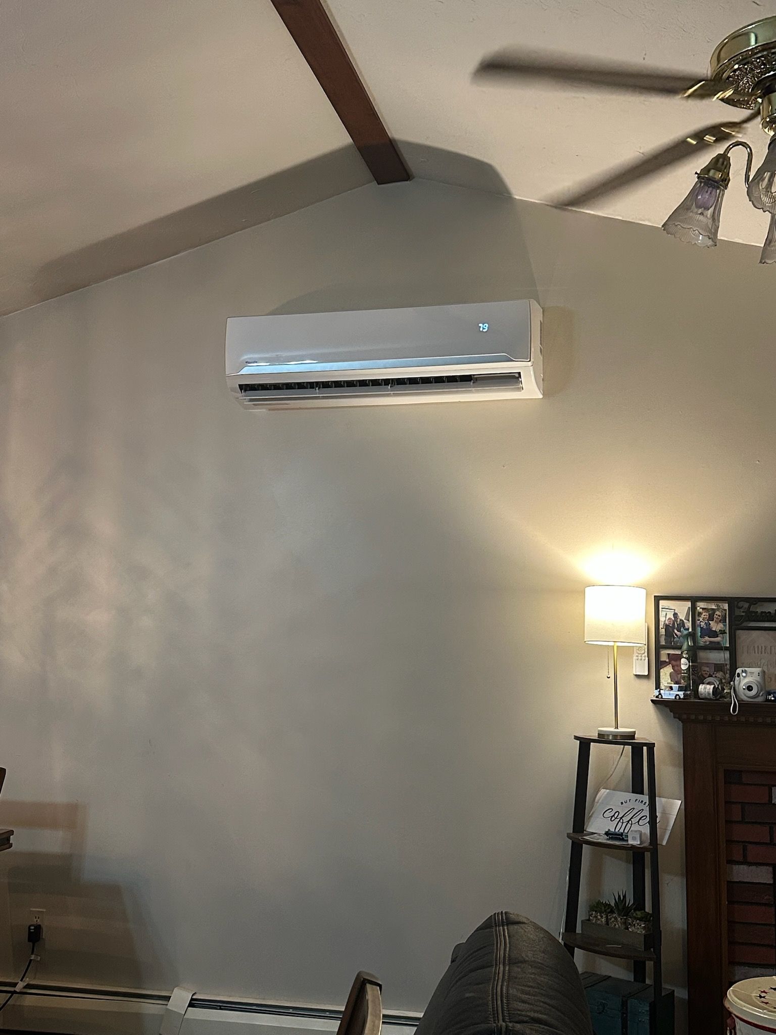 Wall-mounted white air conditioner on a gray wall beneath a vaulted ceiling, with a lit lamp and shelf below.