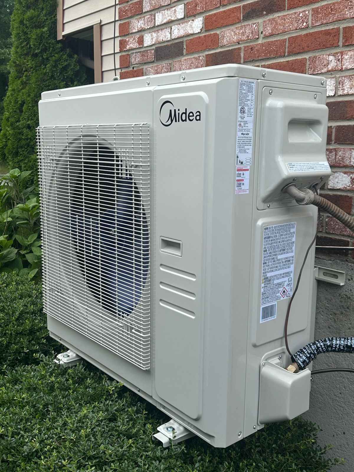 Outdoor Midea wall-mounted air conditioner unit beside a brick house and greenery