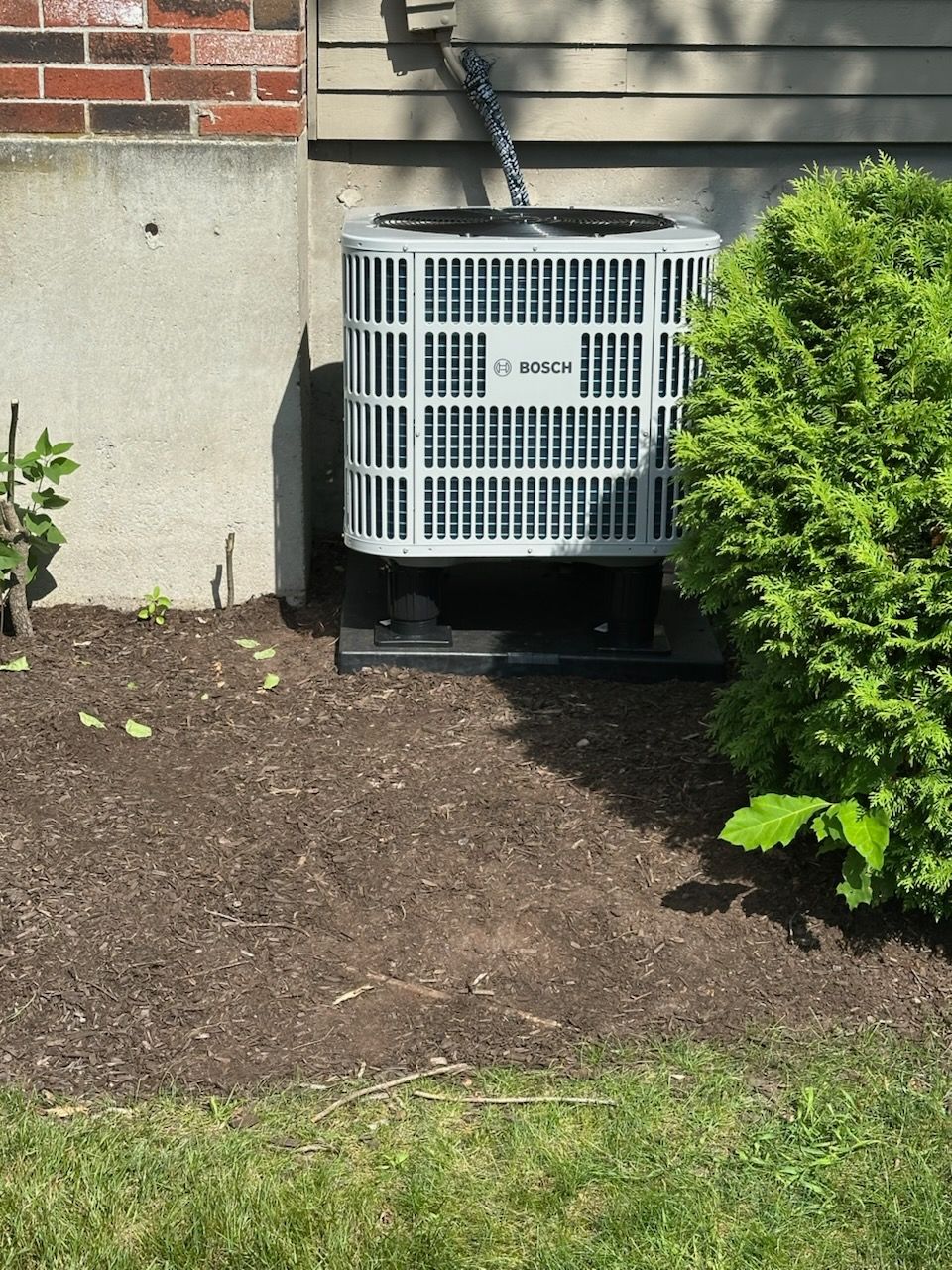 Outdoor air conditioner unit beside a brick wall, surrounded by mulch and grass.