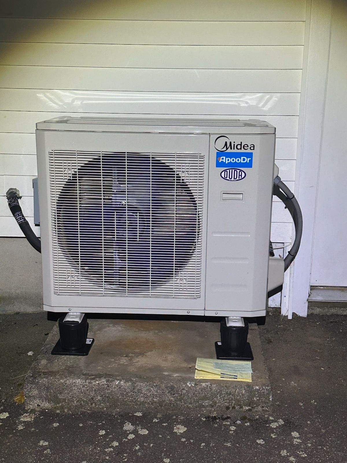 White outdoor air conditioner unit beside a house, with visible fan and hoses on a concrete pad.