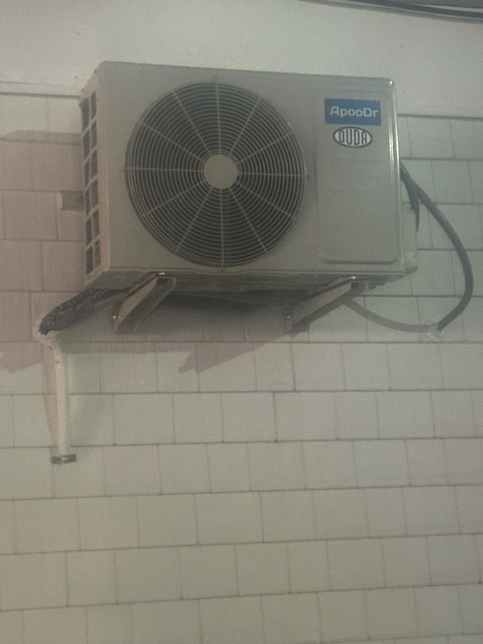 Wall-mounted air conditioner with a large fan unit and dangling cables against a tiled wall.