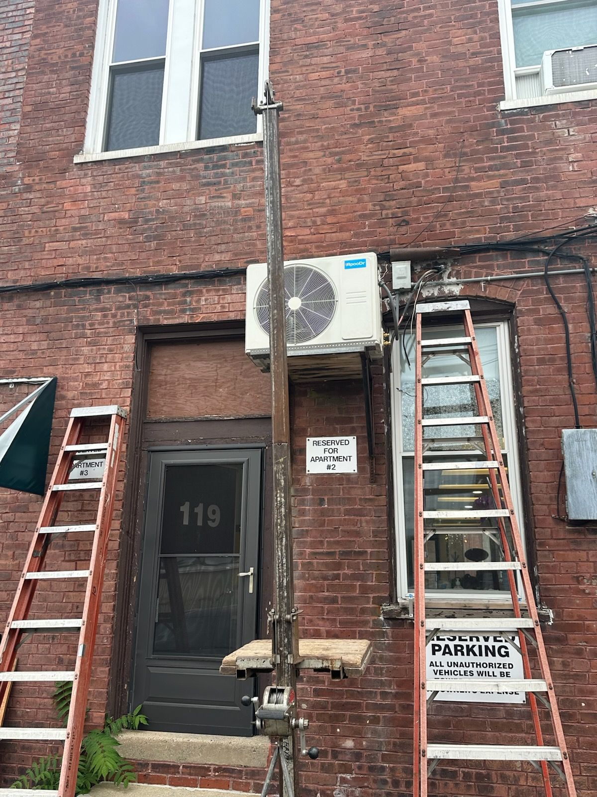 Red brick building entrance with two ladders, an air conditioner unit, and parking signs.