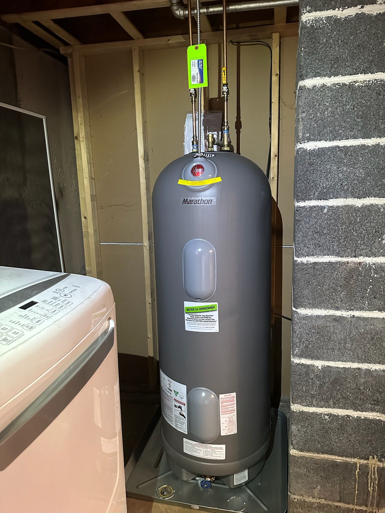 Gray water heater in a utility closet beside a washing machine, with exposed wall framing.