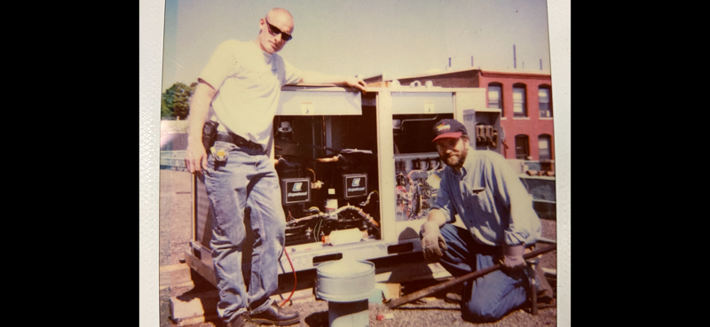 Two workers inspecting equipment on a rooftop, with an open machinery box and a city building in the background.