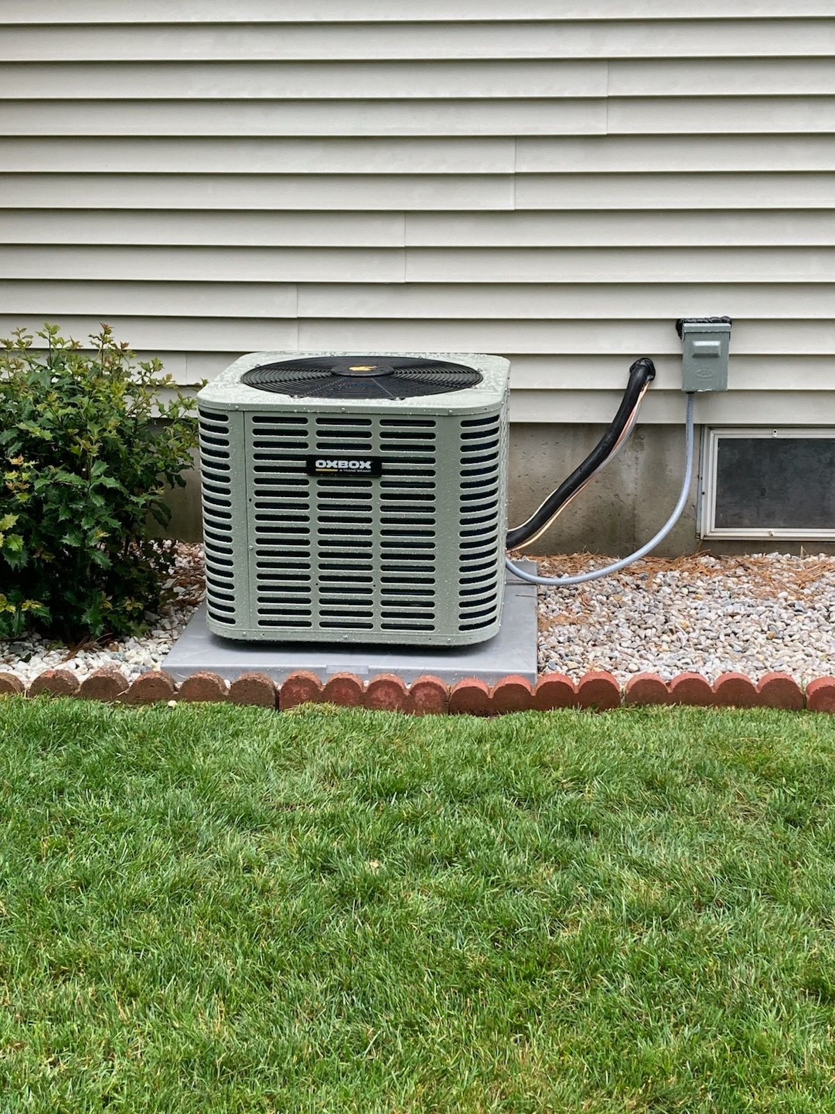 Outdoor air conditioner unit beside a house wall with a hose, gravel border, and green lawn