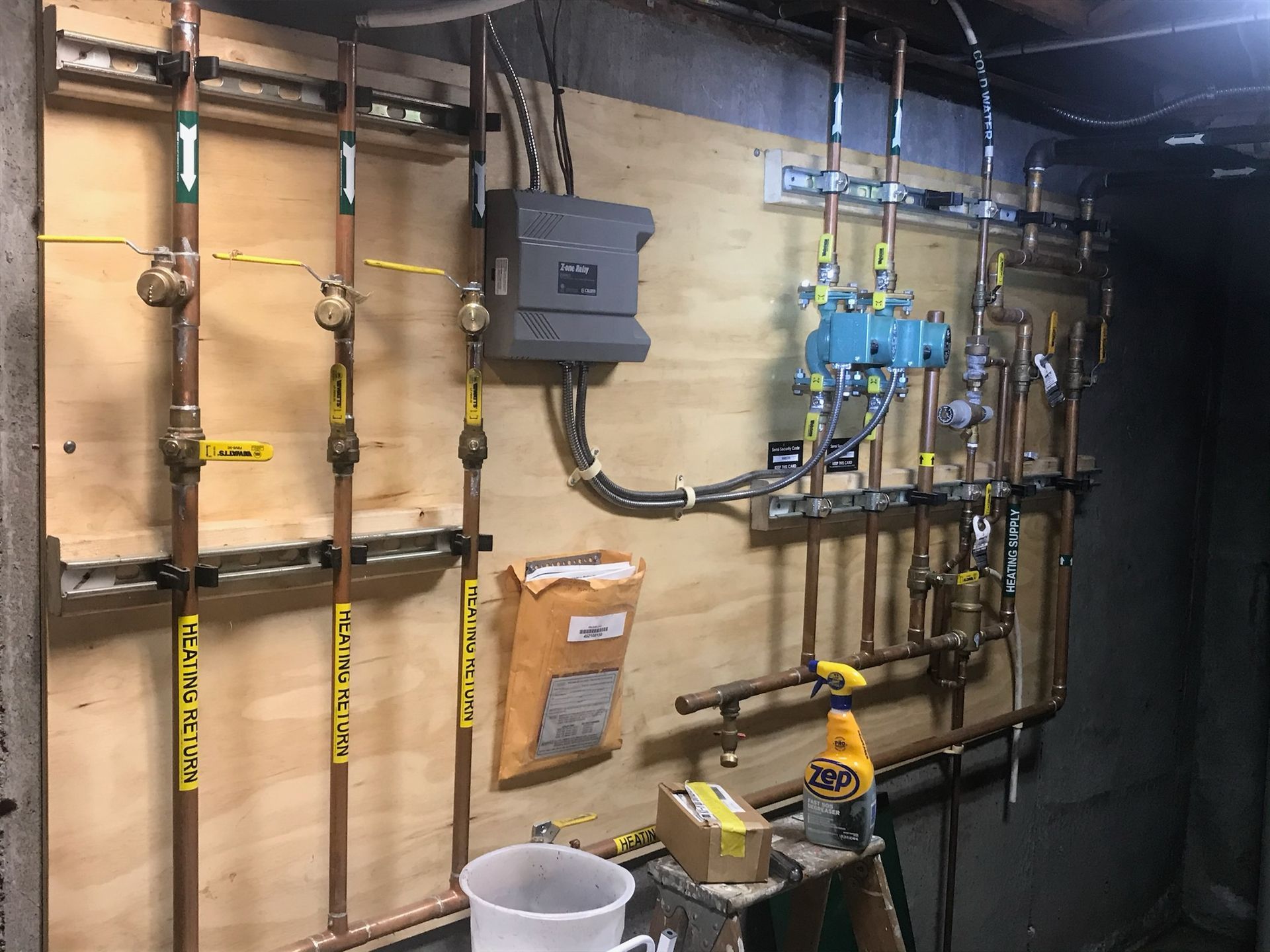 Basement utility wall with pipes, valves, meter box, and a bucket on the floor