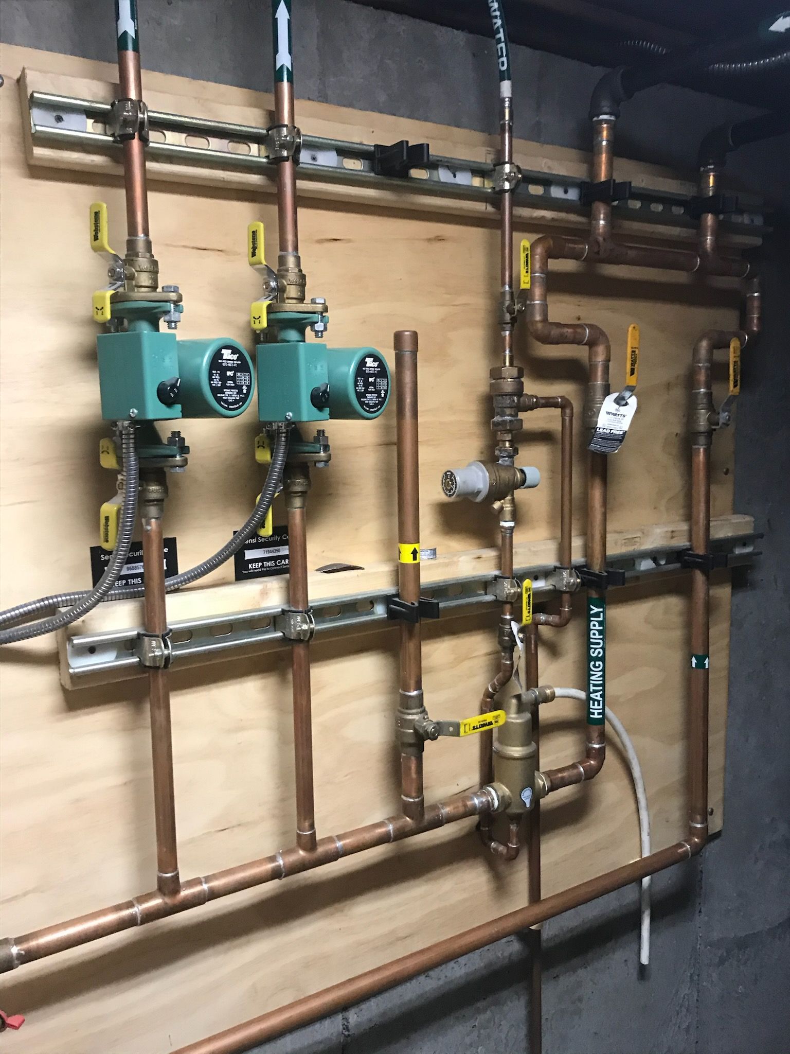 Industrial plumbing manifold with teal pumps, copper pipes, valves, and gauges mounted on a wooden wall.
