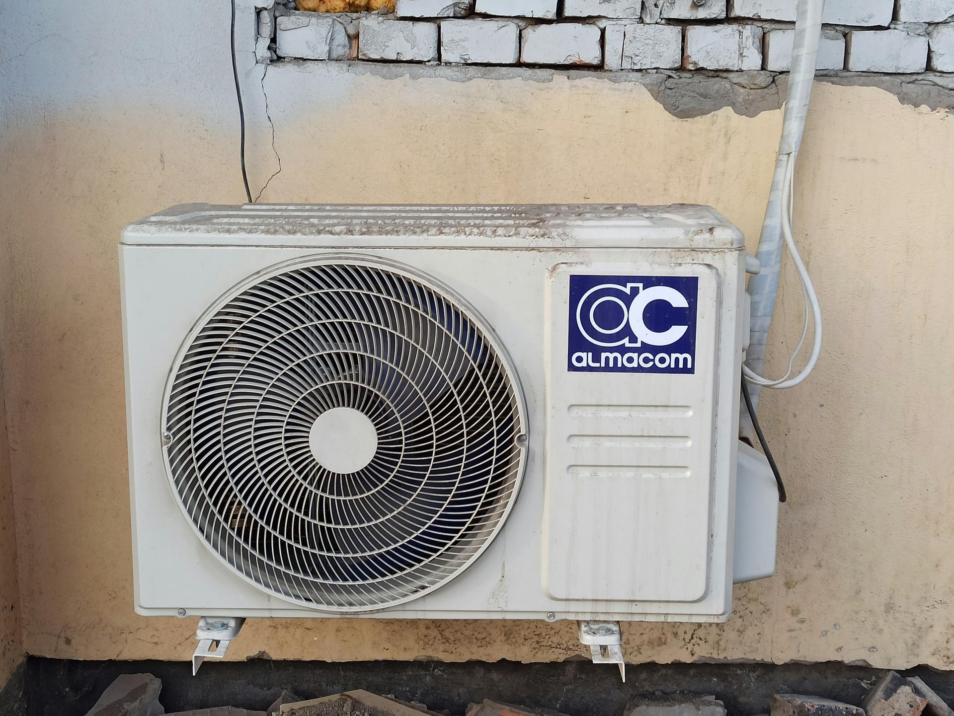 An off-white outdoor air conditioning unit with a circular fan grill mounted on a beige and brick wall.