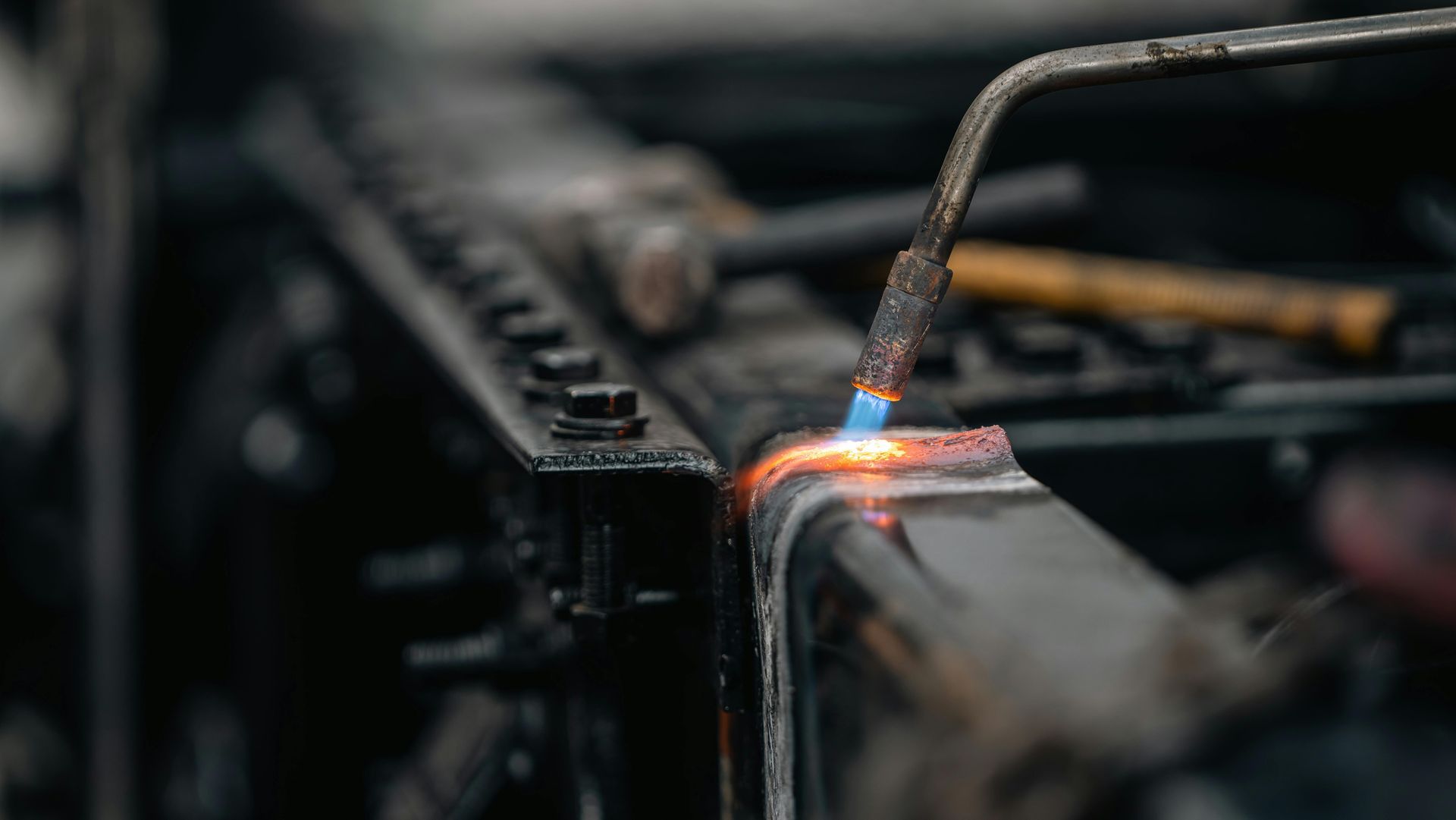 A blowtorch heats a metal frame, causing the surface to glow bright orange against a dark, industrial workshop background.