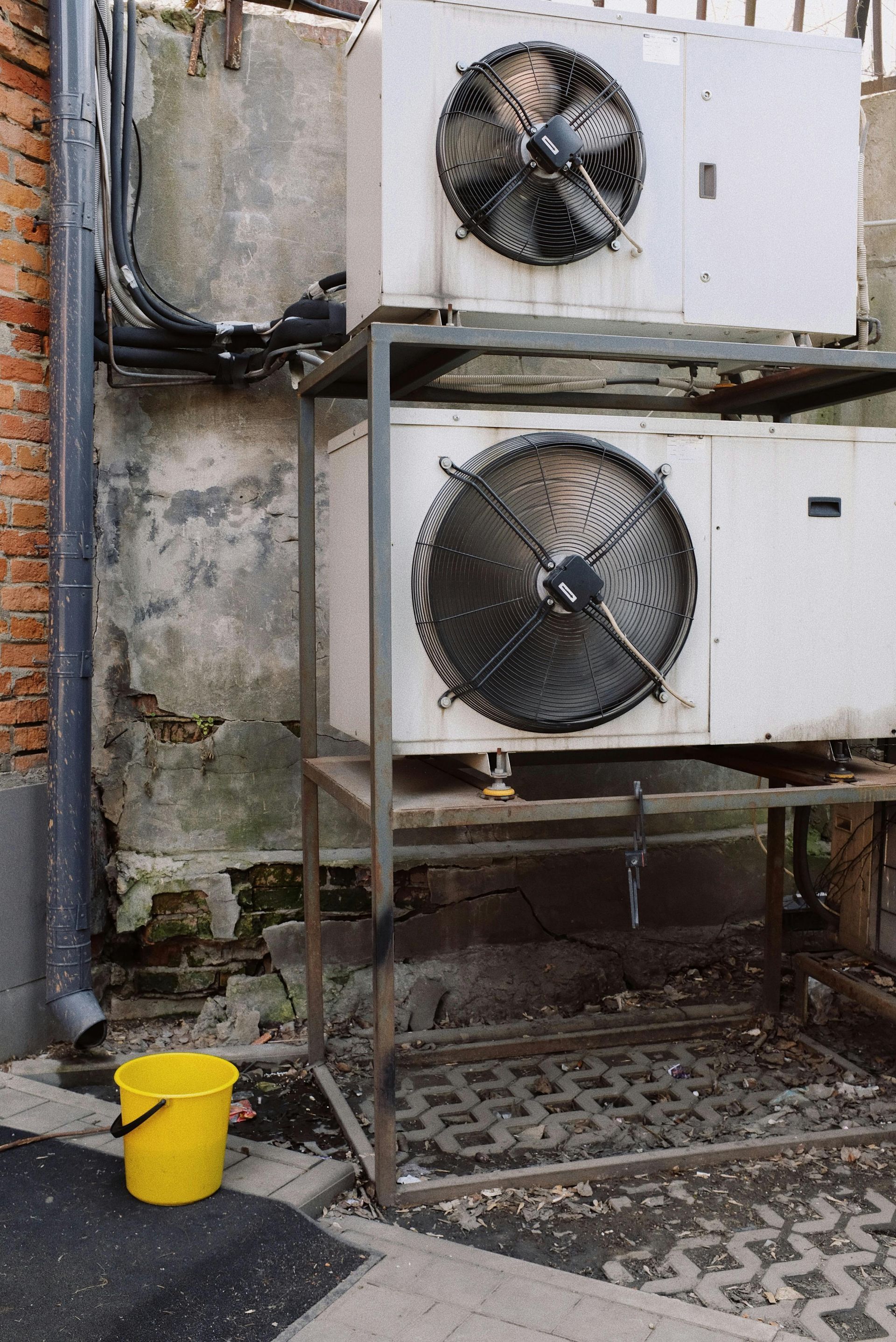 Two stacked industrial air conditioning units on a metal stand beside a weathered brick wall and a small yellow bucket.