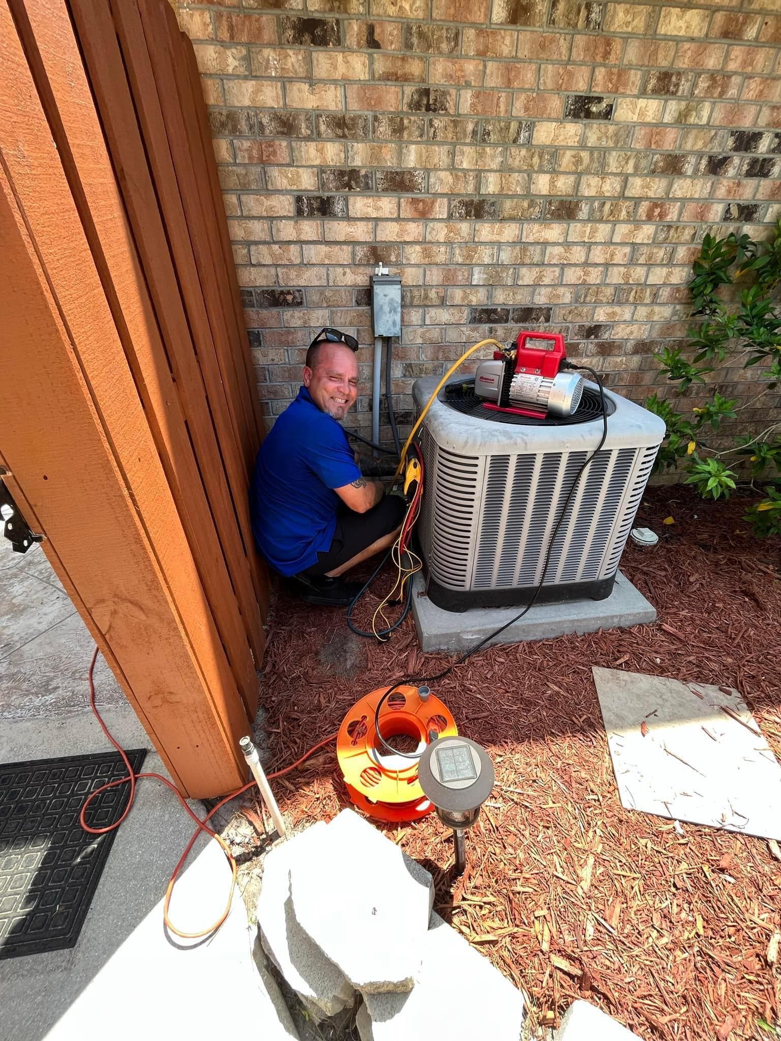 A Local Guy technician is sitting on the ground working on an air conditioner.