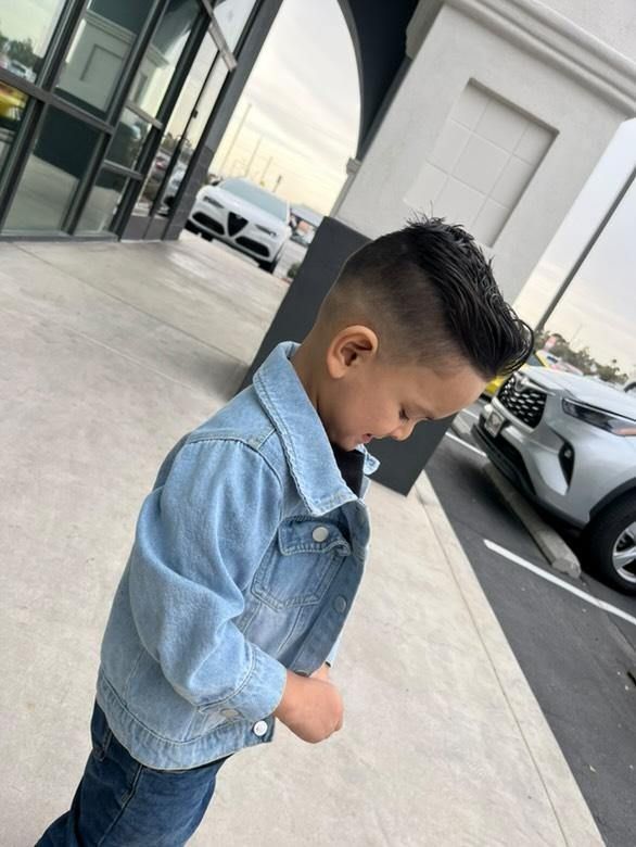 A young child wearing a light blue denim jacket and dark jeans, standing outside on a sidewalk near parked cars.