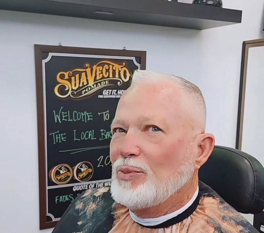 A person with a white beard and a short fade haircut sitting in a barber chair in front of a Suavecito Pomade sign.
