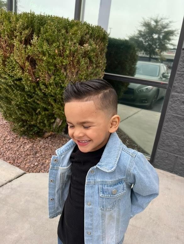 A smiling child with a short fade haircut wears a black shirt and a light-wash denim jacket outdoors.