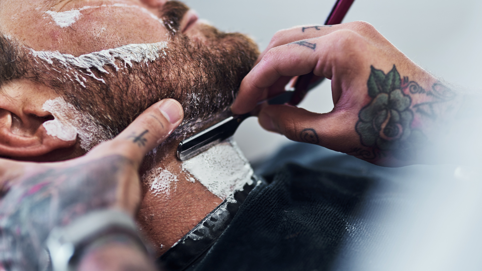 A person with tattooed arms uses a straight razor to shave another person's neck with shaving cream.