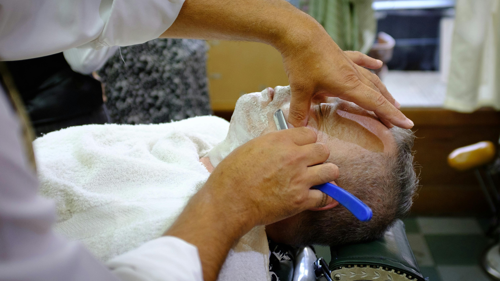 A barber shaves a person’s face using a straight razor with a blue handle, while applying shaving cream.