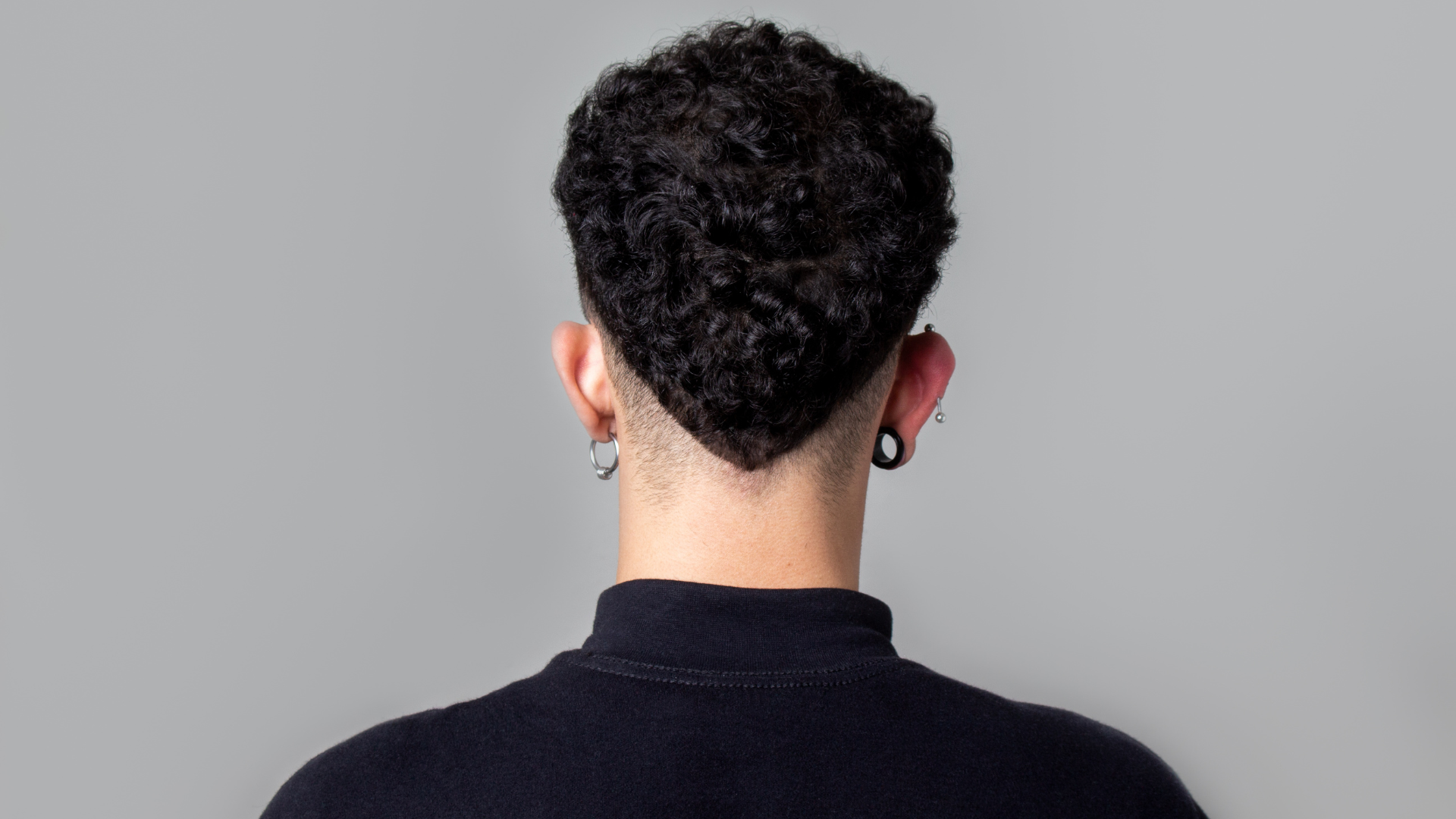 Back view of a person with a short, curly-topped hairstyle that tapers to a V-shape at the nape, wearing dark earrings.