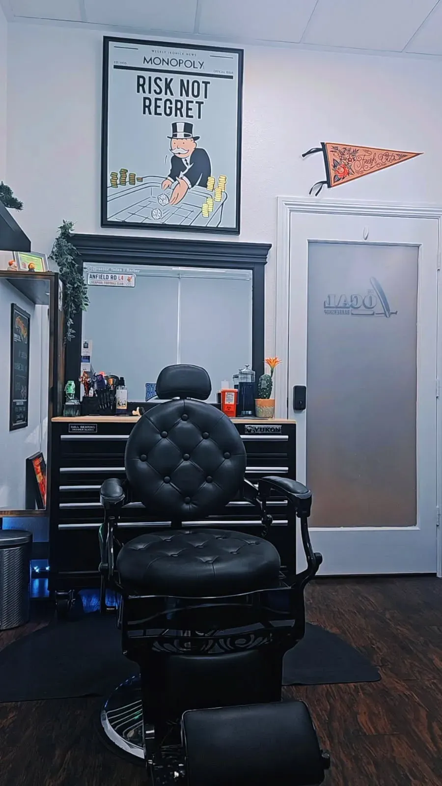 A professional barber chair stands in front of a mirror and a black tool chest in a room with a Monopoly poster on the wall.