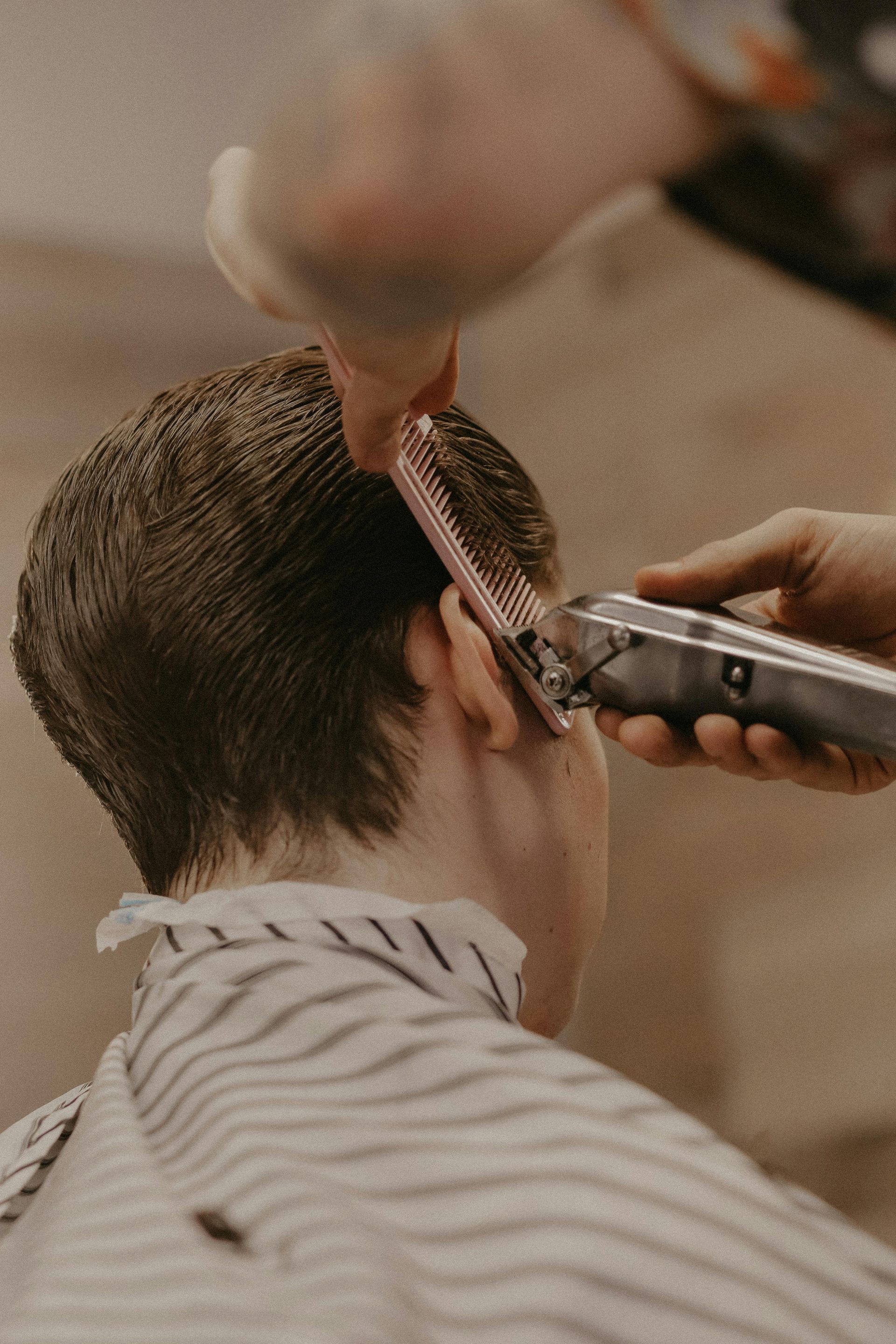 A barber uses electric clippers and a comb to trim hair around a person's ear.